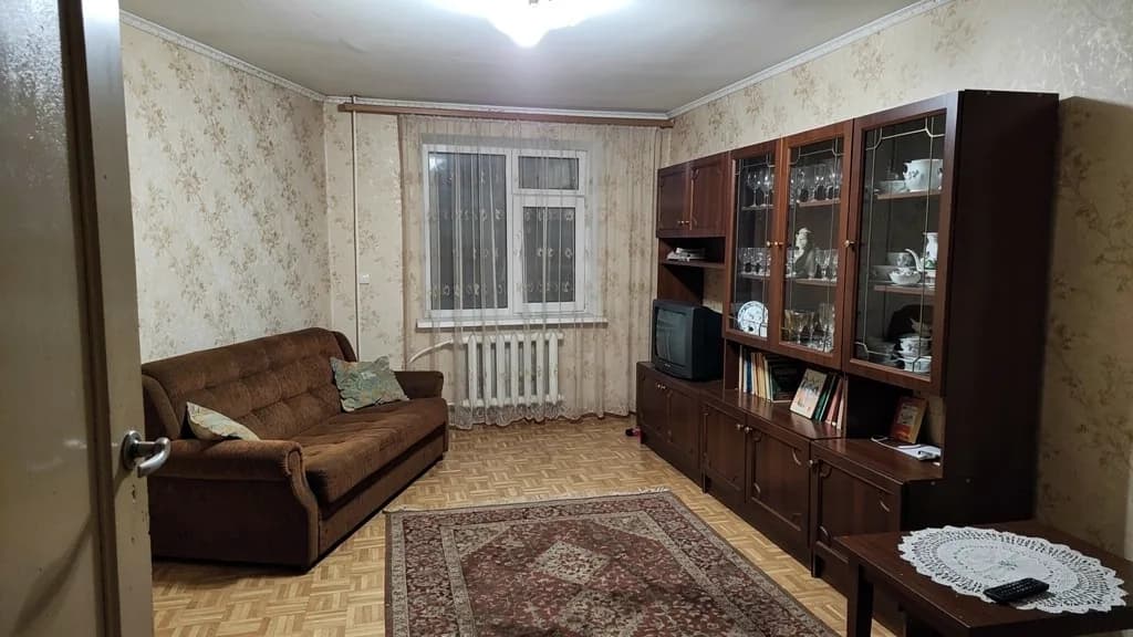 Before: a living room in a standard flat with beige wallpaper, an old sofa and a wall unit, shot on a phone from the door